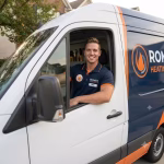 HVAC technician smiling from the driver's seat of a Roman Heating & Air service van parked in a residential neighborhood.